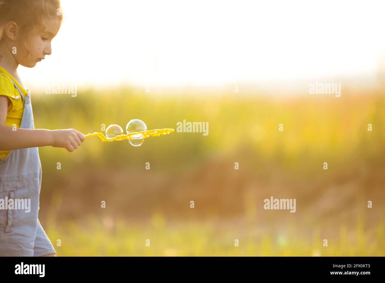 A girl in a denim jumpsuit blows soap bubbles in the summer in a field at sunset. International Children's Day, happy child, outdoor activities. Summe Stock Photo