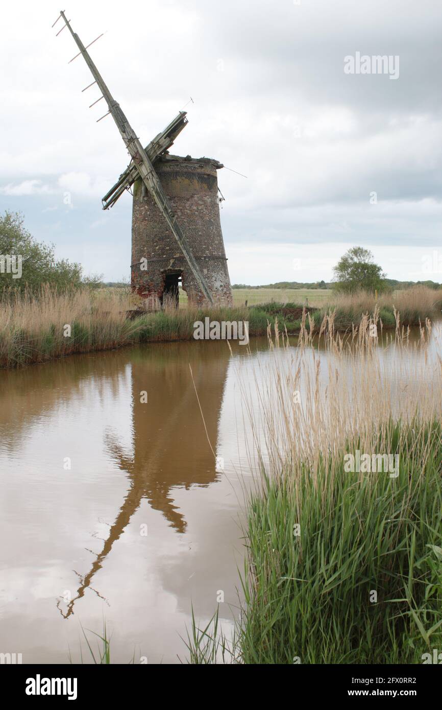 Landscape of ancient brick windmill mill pump with wood sails reflect in river water of Norfolk Broads Horsey East Anglia England by grassy reed bank Stock Photo