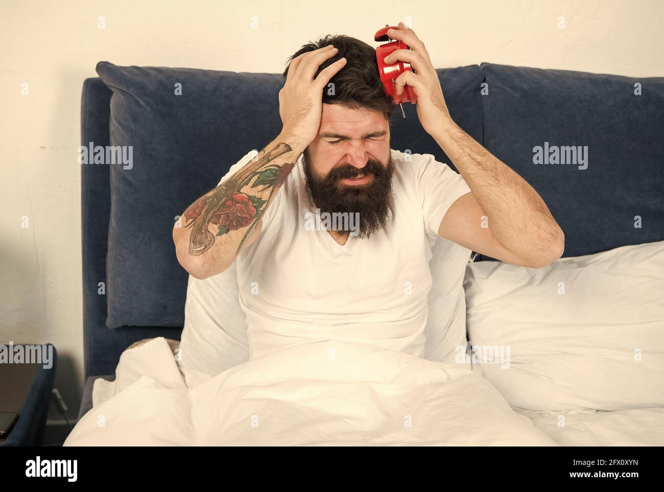 My clock is running late. Late riser suffer headache. Stressed hipster hold alarm clock in bed. Bearded man wake up late in morning. Keeping late Stock Photo