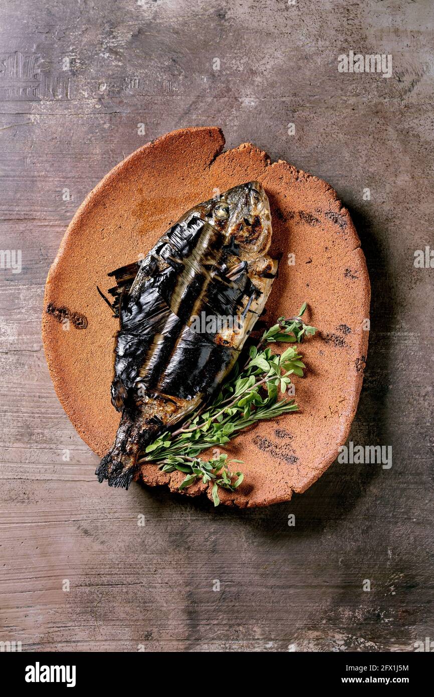 Grilled cooked fresh gutted sea bream or dorado fish on ceramic plate wrapped in bamboo leaves served with herbs over dark brown texture background. T Stock Photo