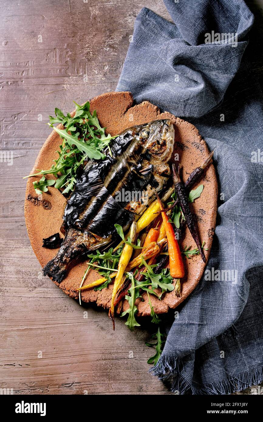 Grilled cooked fresh gutted sea bream or dorado fish on ceramic plate wrapped in bamboo leaves served with herbs, colorful carrots, blue napkin over d Stock Photo