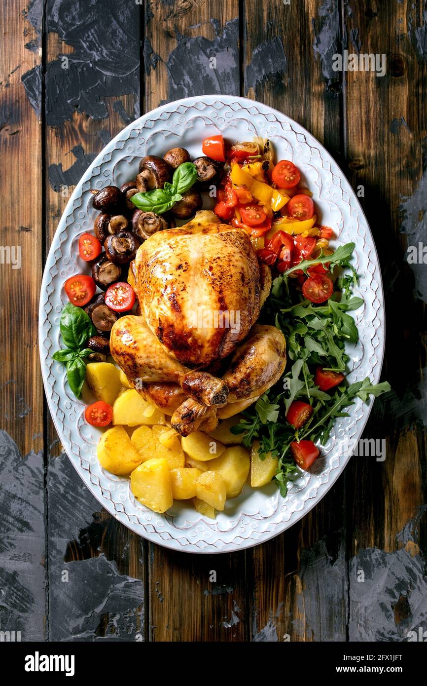 Grilled cooked whole chicken with vegetable garnish grilled bell pepper, onion, baked potatoes, cherry tomatoes, mushrooms and herbs in ceramic plate Stock Photo