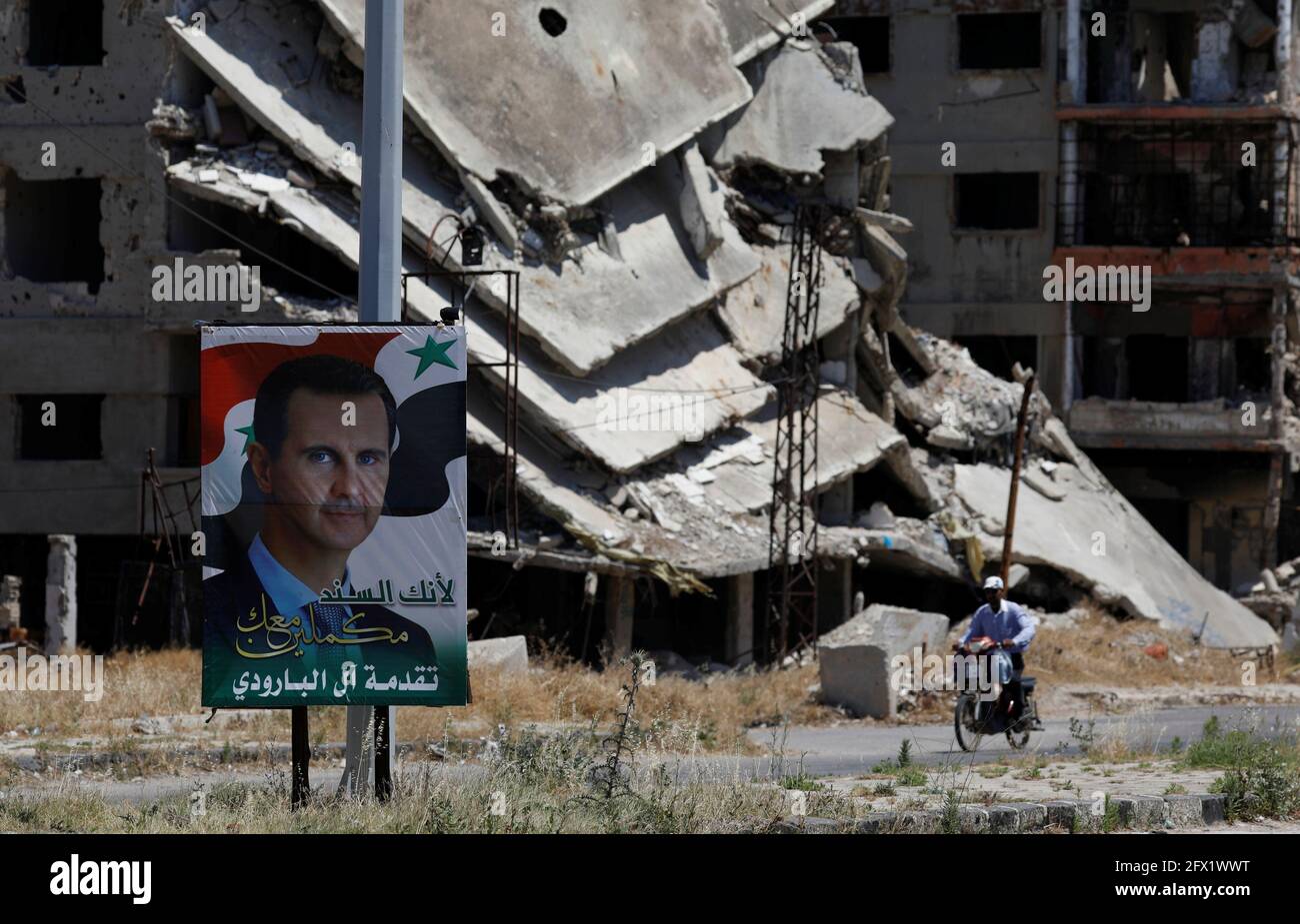 A poster depciting Syria's President Bashar al-Assad is pictured in front of damaged buildings, ahead of the May 26 presidential election, in Homs, Syria May 23, 2021.  Picture taken May 23, 2021. REUTERS/Omar Sanadiki Stock Photo