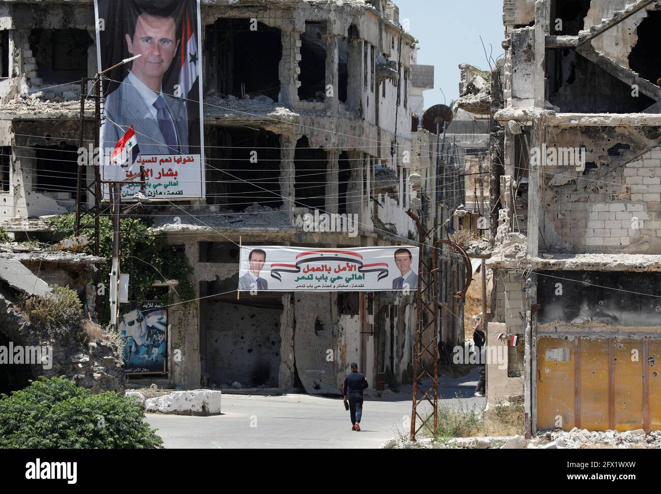 A man walk past banners depciting Syria's President Bashar al-Assad, near damaged buildings, ahead of the May 26 presidential election, in Homs, Syria May 23, 2021.  Picture taken May 23, 2021. REUTERS/Omar Sanadiki Stock Photo