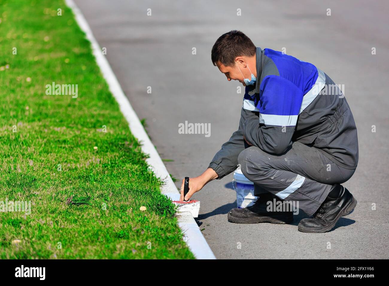 A worker in overalls paints the curb with white paint on a summer day. City service, improvement of territories. A young contractor of Caucasian appearance at work. Stock Photo