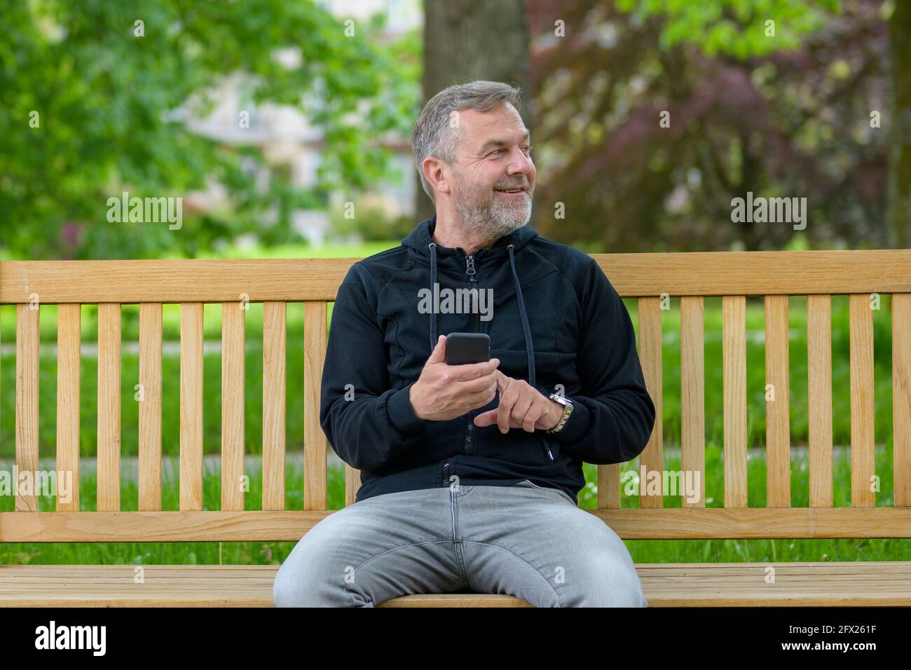 Happy relaxed middle-aged man using his mobile phone in a park texting a message while looking to the side Stock Photo