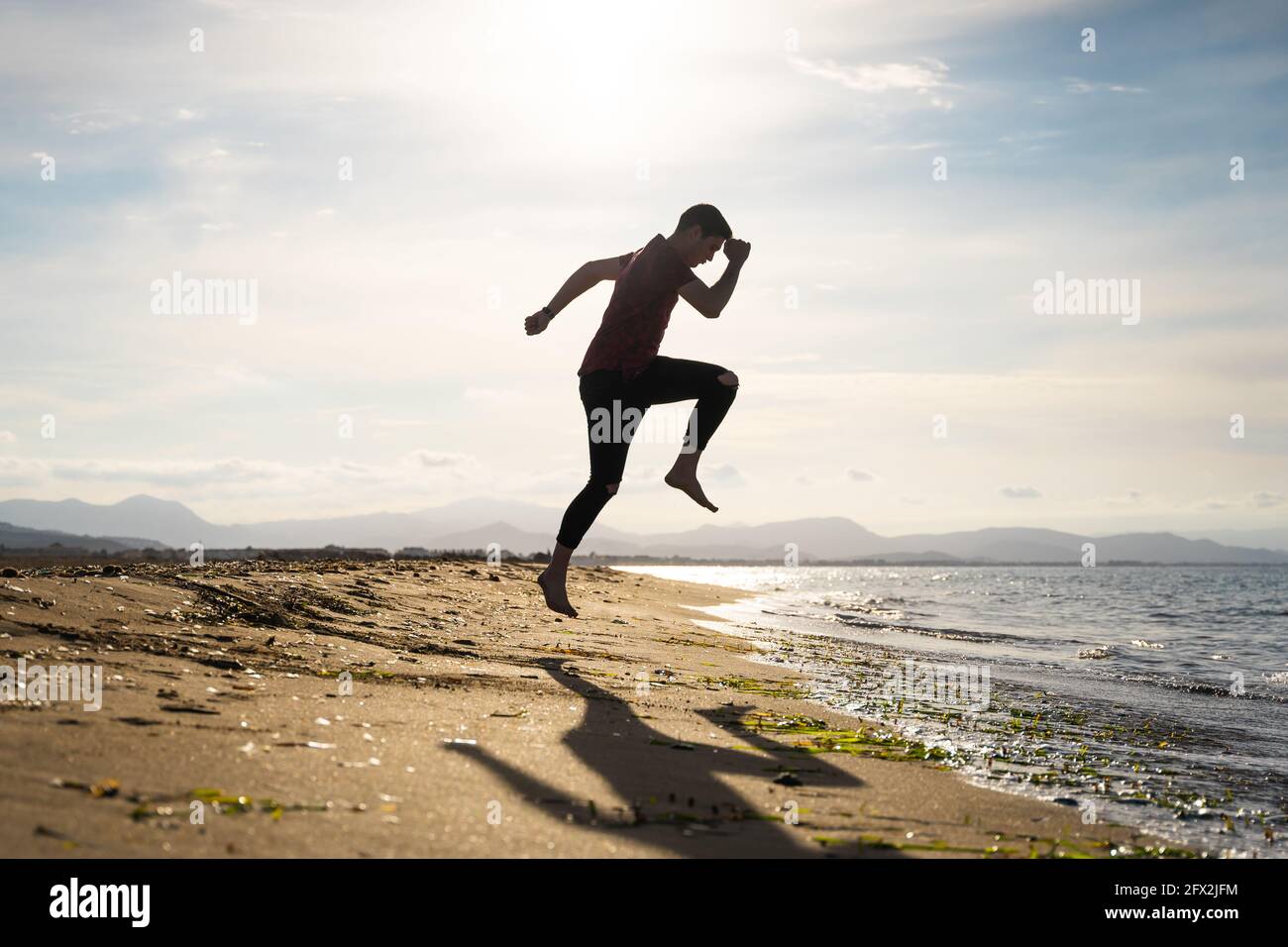 Man jumping on the seashore on a sunny day. Caucasian with long trousers and short-sleeved shirt. Shadow can be seen on the golden sand. Stock Photo