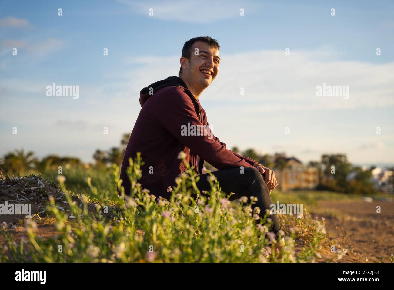 Happy man sitting on the beach smiling. He is wearing a red sweatshirt and black trousers. Caucasian Stock Photo