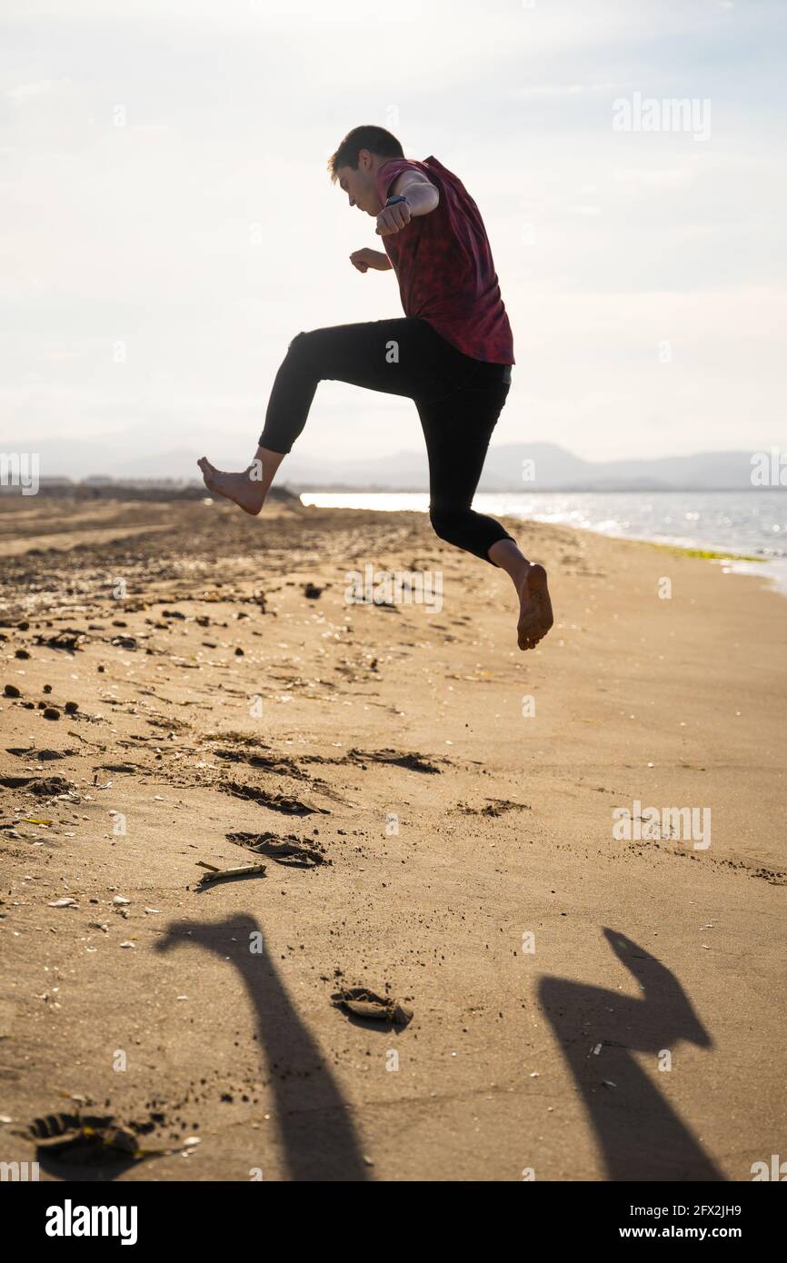 Man jumping on the seashore on a sunny day. Caucasian with long trousers and short-sleeved shirt. Shadow can be seen on the golden sand. Stock Photo