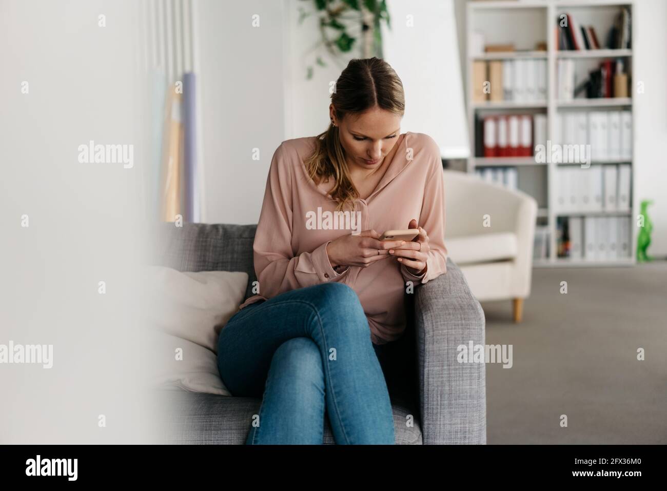 Young woman reading with concentration a message or browsing the internet on the mobile phone while sitting on a sofa in a modern office Stock Photo