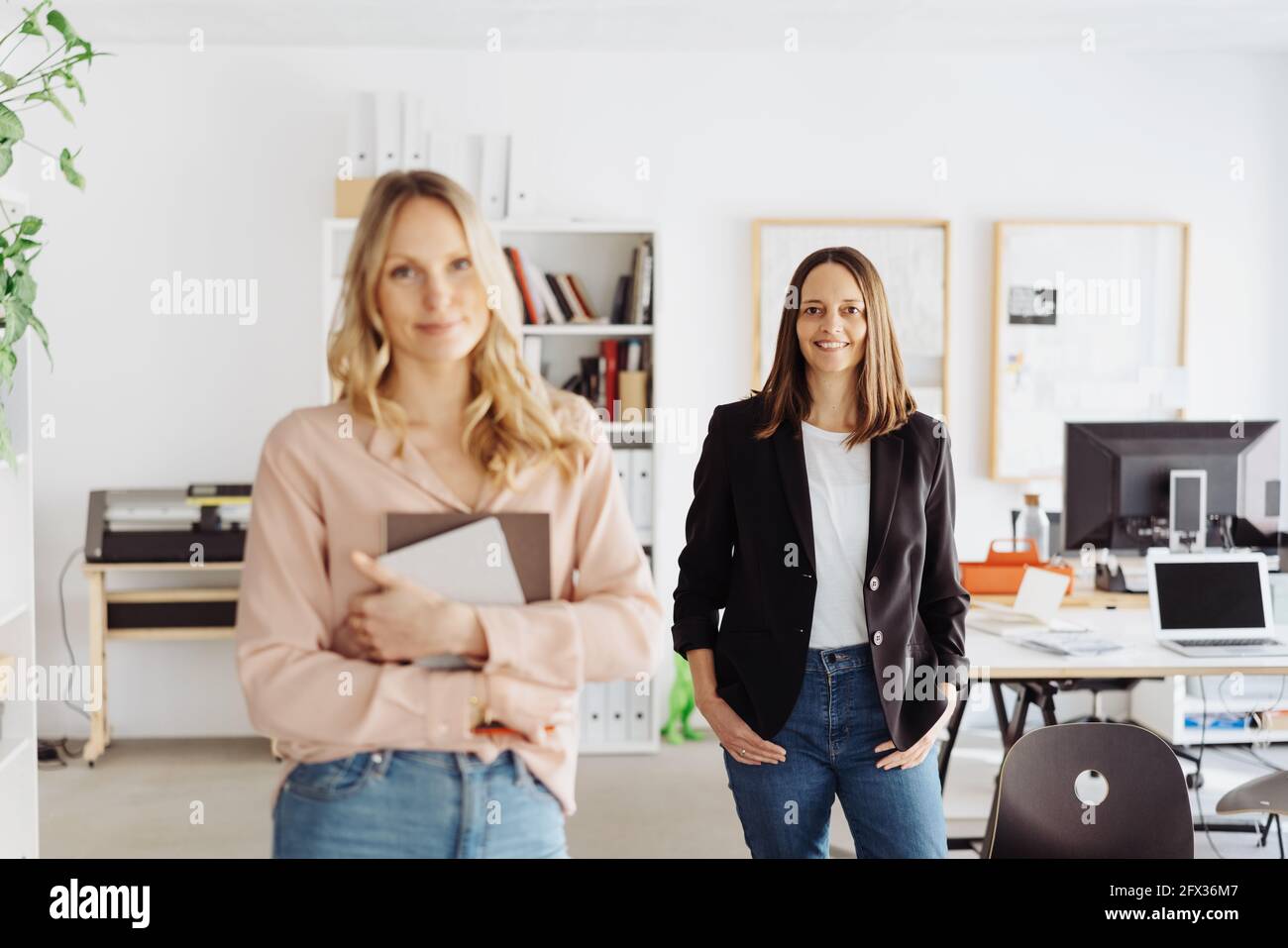 Two confident successful female business partners or colleagues posing in a spacious modern office with focus to the smart woman in the rear smiling a Stock Photo