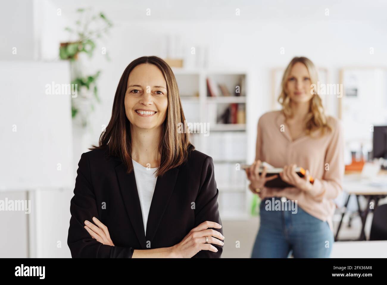 Happy confident successful manageress or business partner posing with folded arms smiling at the camera with her colleague behind her Stock Photo
