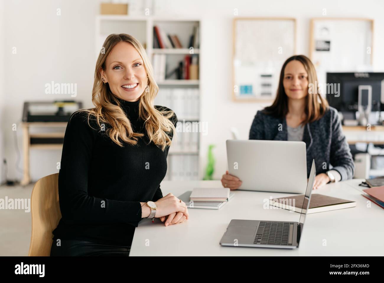 Smiling friendly sincere businesswoman turning to look at the camera as she sits at an office table with a colleague working in the background in a hi Stock Photo