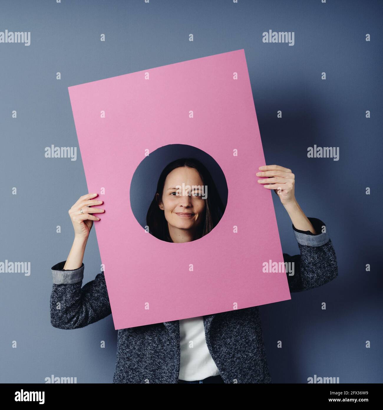 Smart middle-aged woman framing her face with a pink placard with central circular cutout looking at the camera with a friendly smile over a blue stud Stock Photo