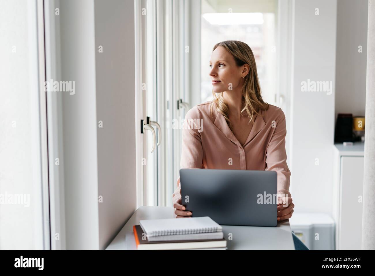 Young woman sitting at a laptop computer daydreaming looking out of the window alongside with a contemplative faraway expression in a high key office Stock Photo