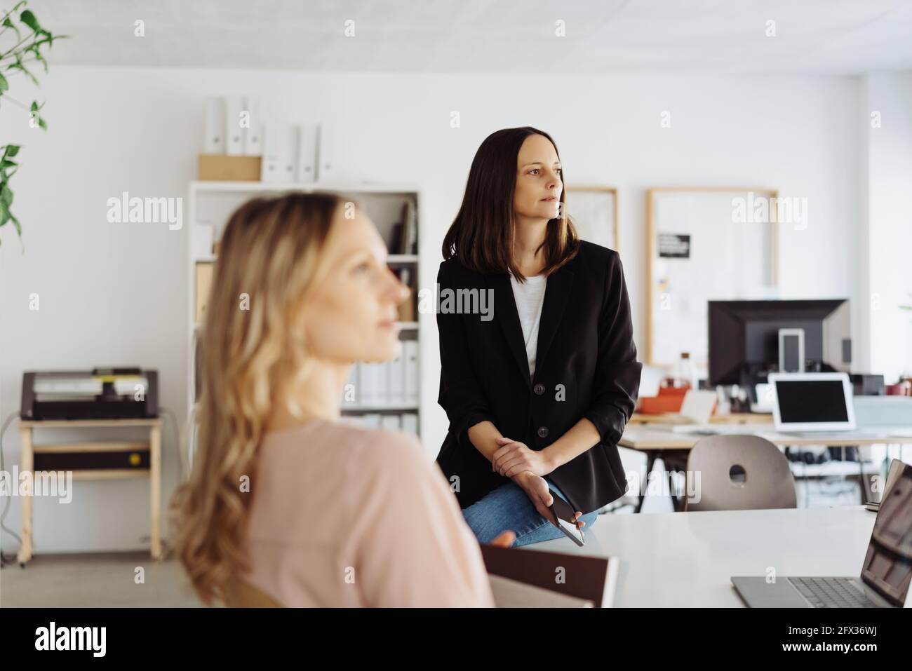 Two businesswoman sitting in a meeting looking to the side listening with focus to the woman in the rear perched on the edge of the table in a high ke Stock Photo