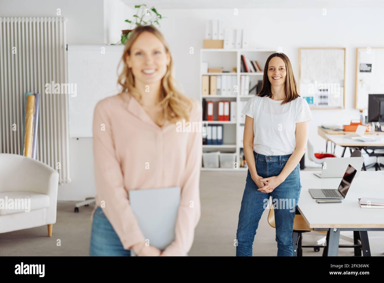 Two work colleagues or business partners posing in a high key modern open plan office with focus to the woman in the rear in casual jeans smiling at t Stock Photo