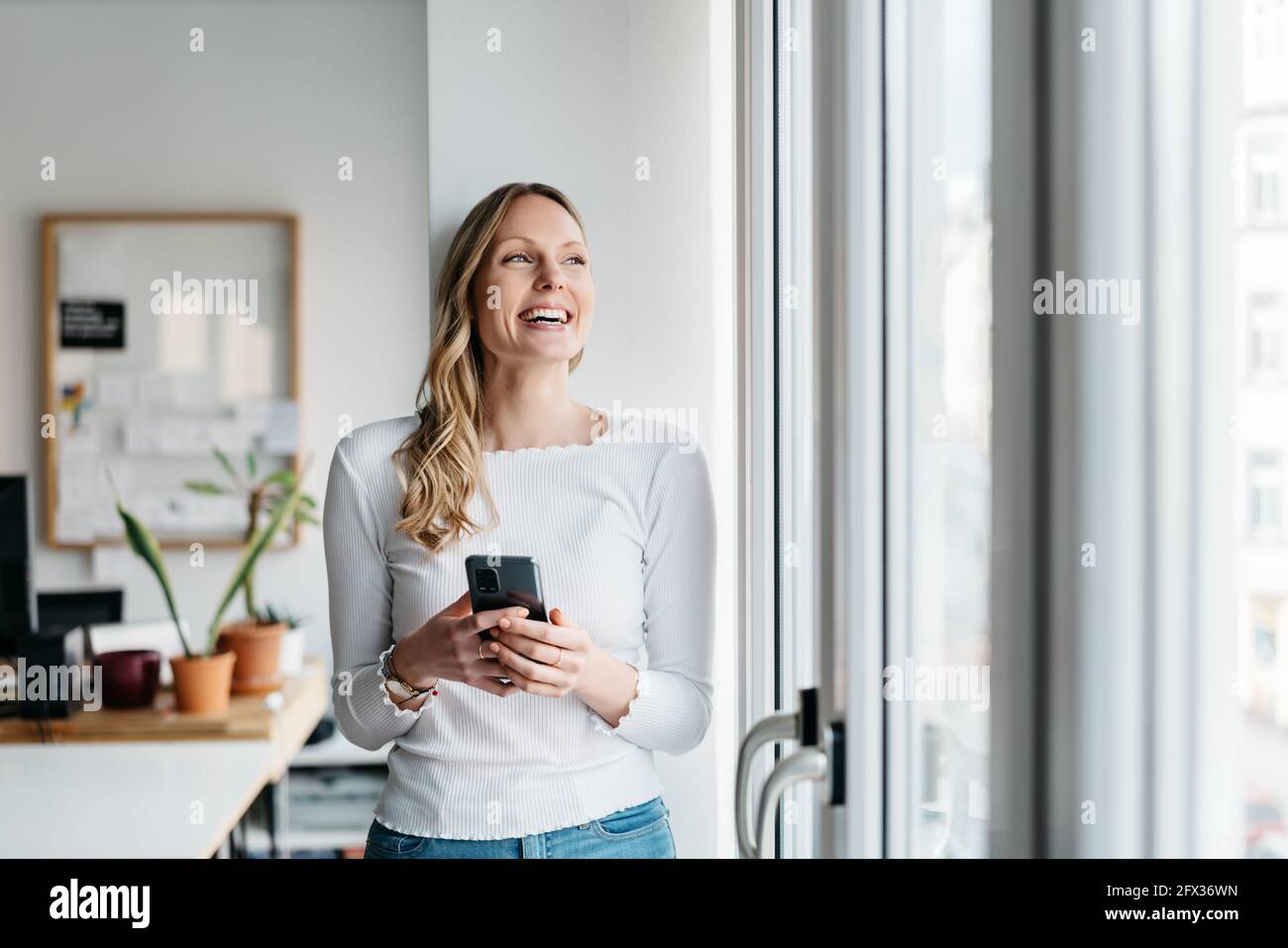 Young woman standing in the office looking out through a glass door with a beaming smile of pleasure as she holds her mobile phone in her hands Stock Photo