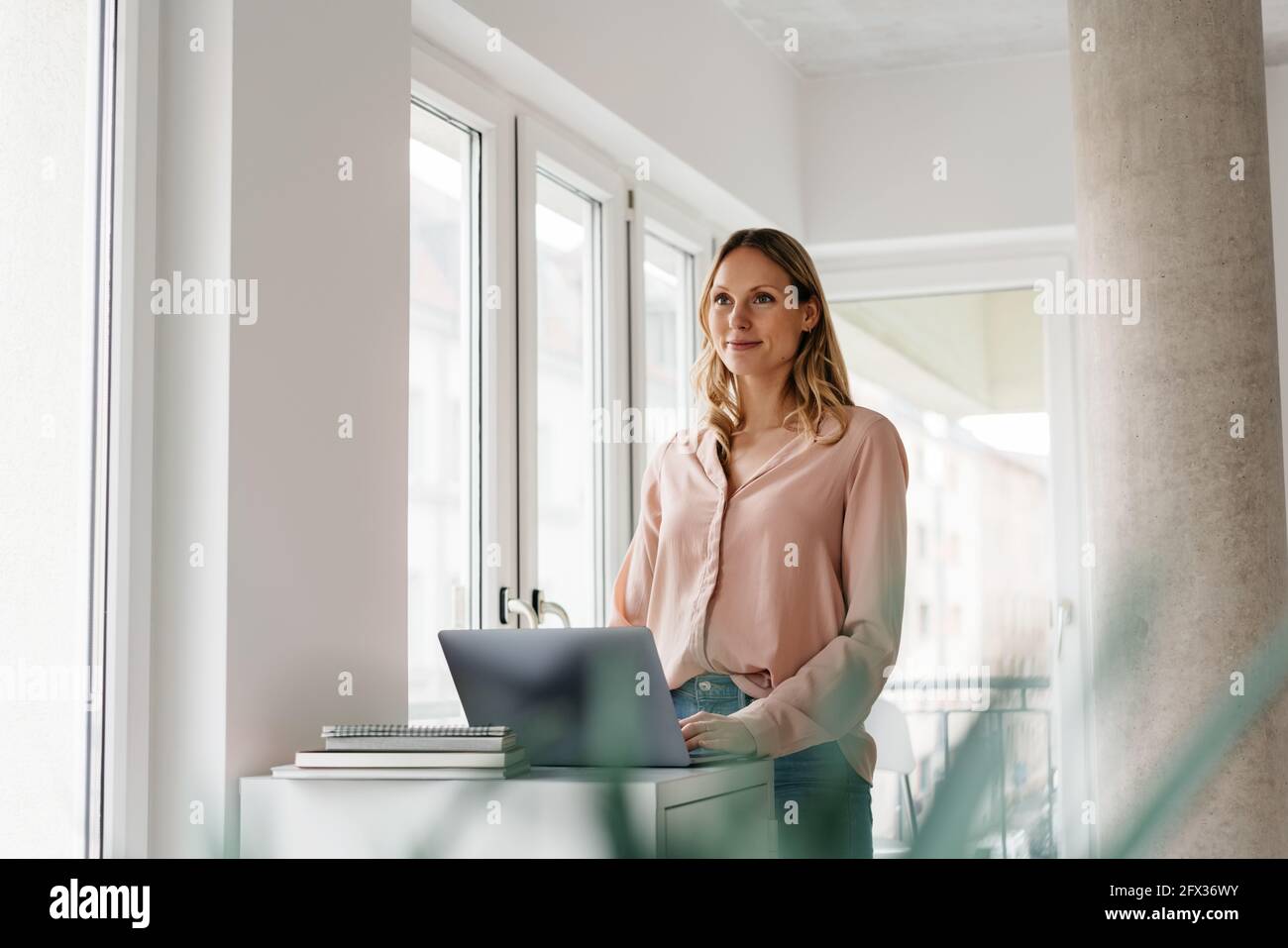Young woman working at a laptop standing staring ahead with a thoughtful smile in a bright high key office as she works from home during the Covid-19 Stock Photo