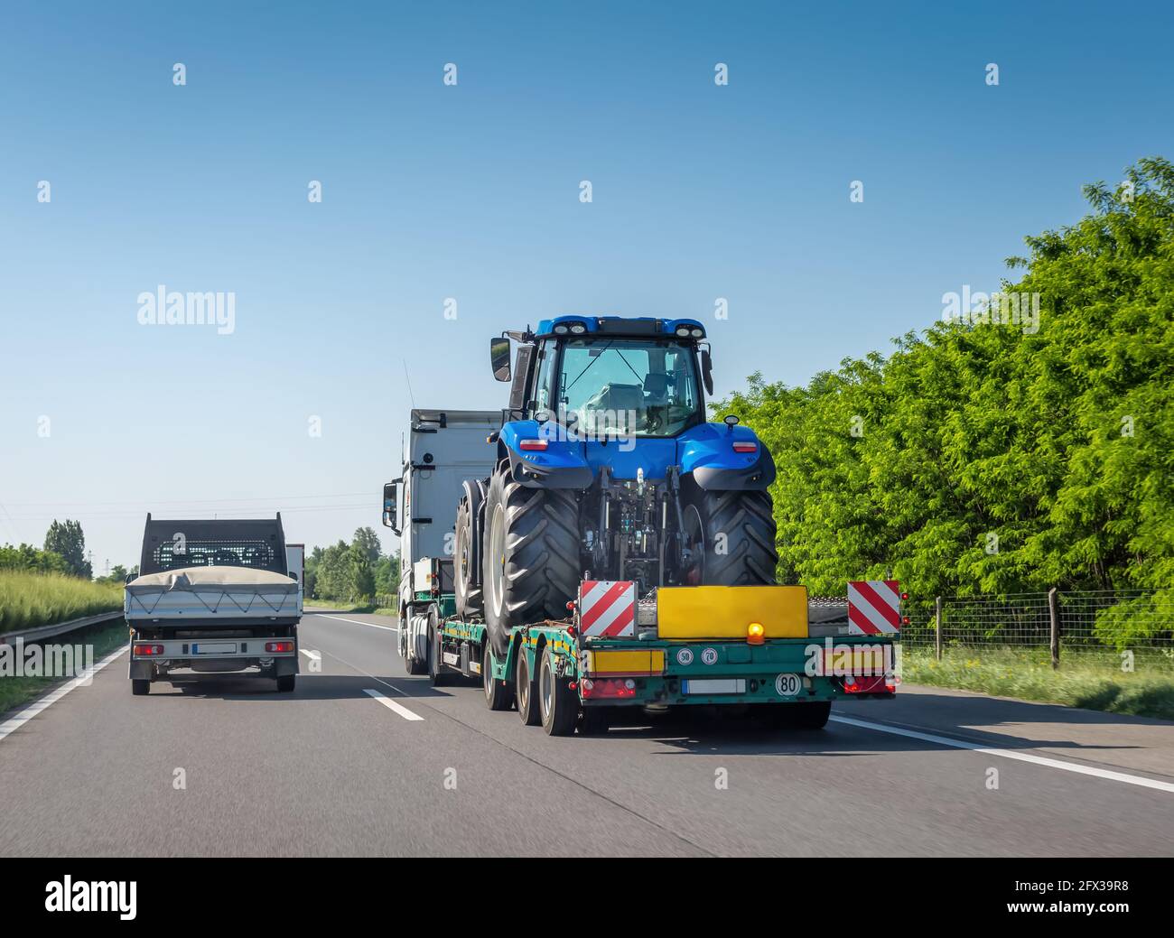 New blue tractor on long trailer platform of truck for transporting heavy machinery on highway. Transportation of agricultural machinery Stock Photo