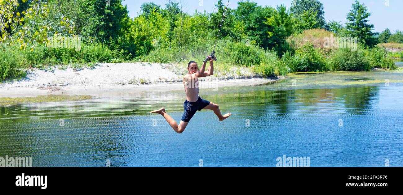 Boy teenager swings on a rope in a funny pose and ready to jump into the water on sunny summer day. Beautiful landscape of a river with green banks. C Stock Photo