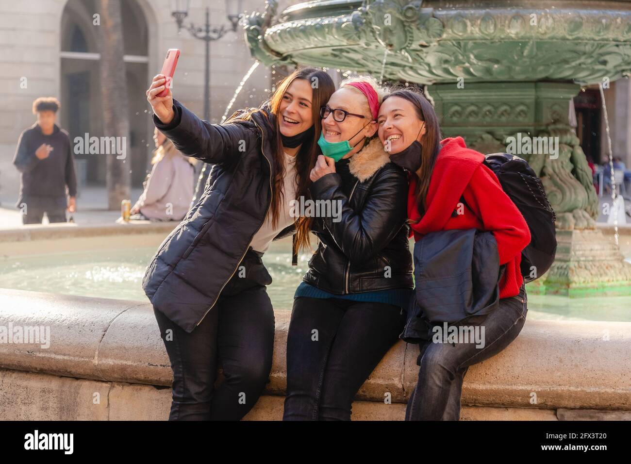 Three women friends capturing a selfie with a smart phone during covid 19 pandemic, wearing protective face masks, in centre of Barcelona Stock Photo