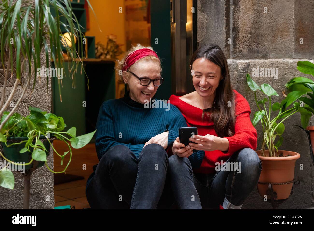 Two middle-aged women friends sitting close together with smart phone having fun in front of a shop. Concept: female friendship, closeness, everyday u Stock Photo