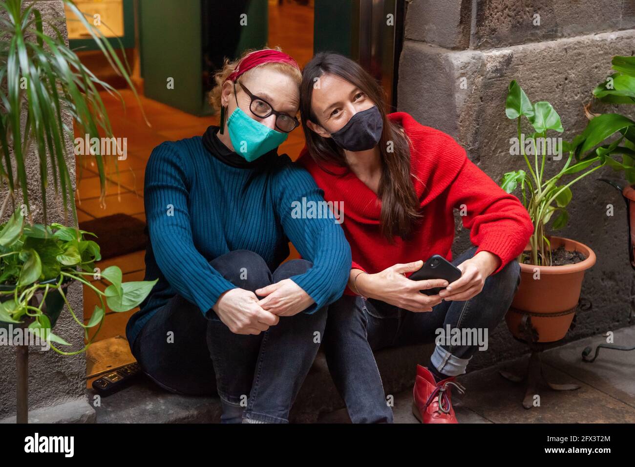 Portrait of two middle-aged women friends with protective face masks sitting close together in the entrance of their shop. Concept: working during cor Stock Photo