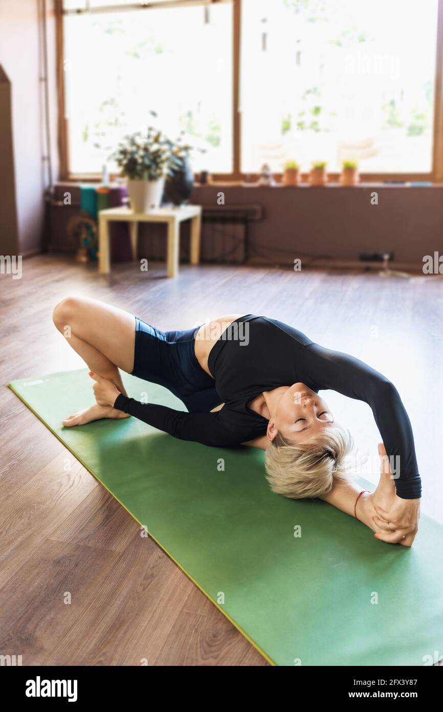A beautiful woman in sportswear, practicing yoga, performs the parivritta janu shirshasana exercise on the rug near the window, tilting her head to th Stock Photo