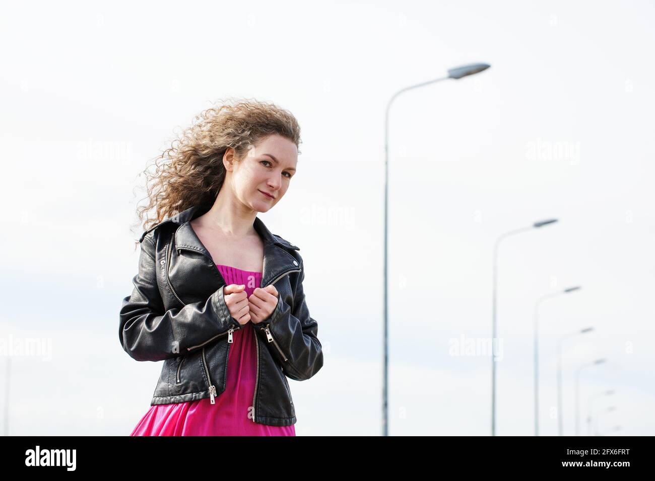 caucasian curly-haired woman in a black leather jacket and a pink dress against the background of a cloudy sky and pillars with lanterns Stock Photo