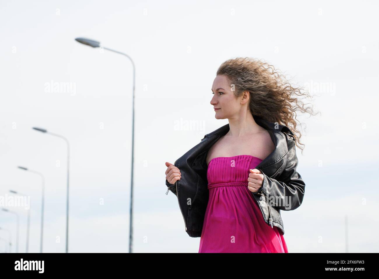 caucasian curly-haired woman in a black leather jacket and a pink dress against the background of a cloudy sky and pillars with lanterns Stock Photo