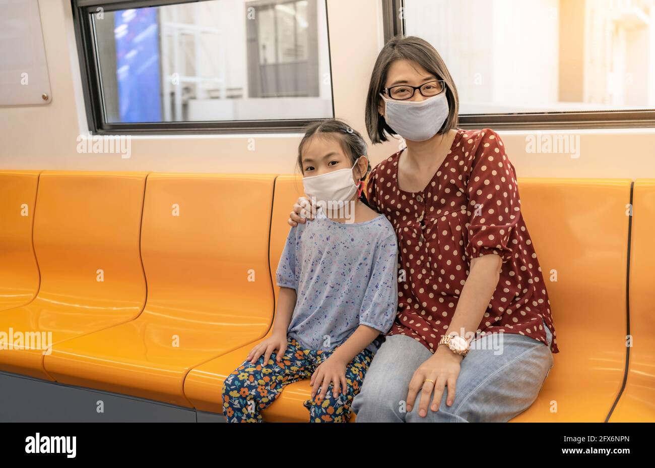 Portrait of Asian or Thai mother and little daughter sitting on sky train of Bangkok, Thailand. Mother and little daughter wearing protective mask whi Stock Photo