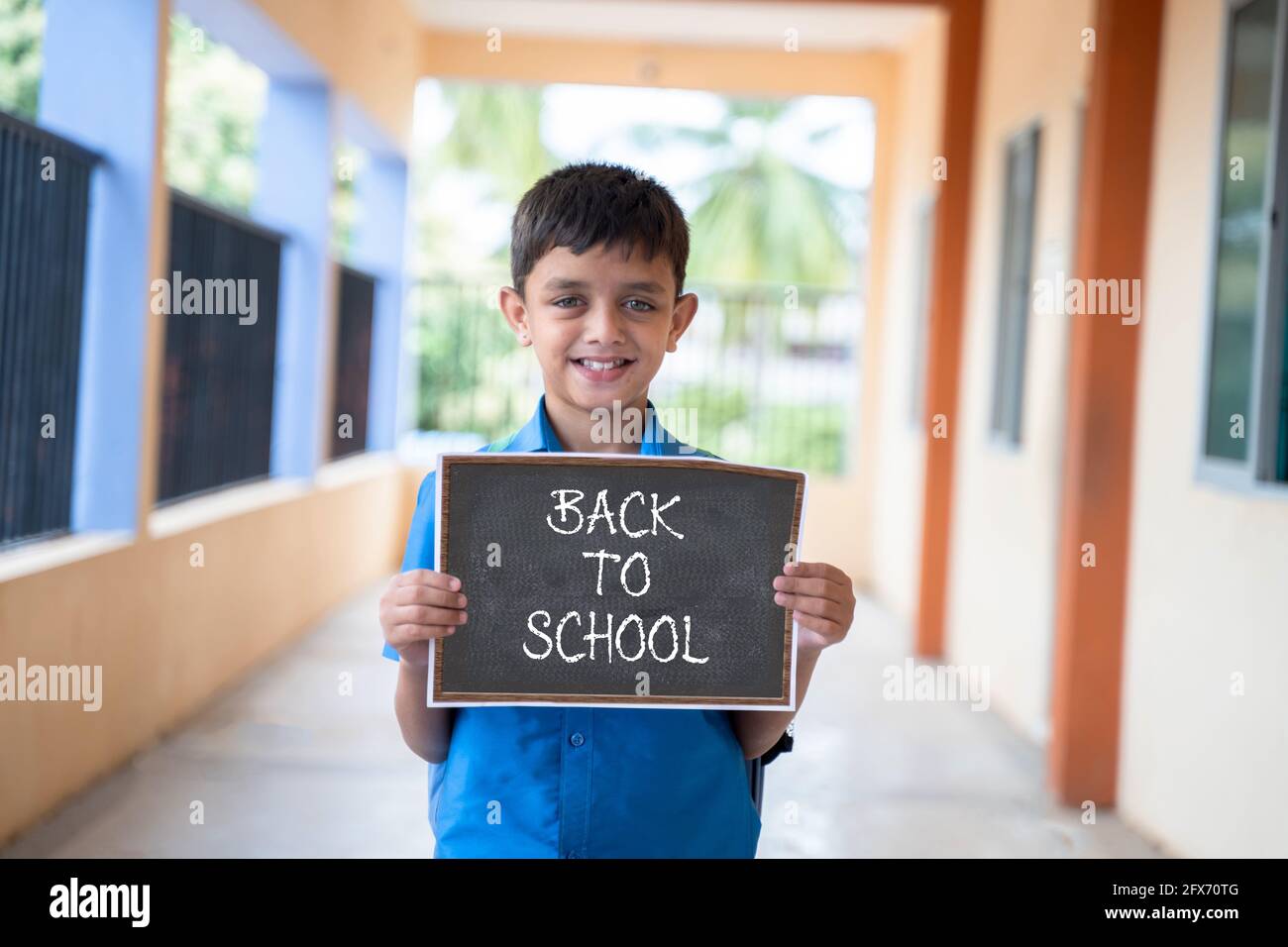 Smiling Young kid looking camera and showing by back to school sign board school at corridor - concept of school reopen and back to school. Stock Photo