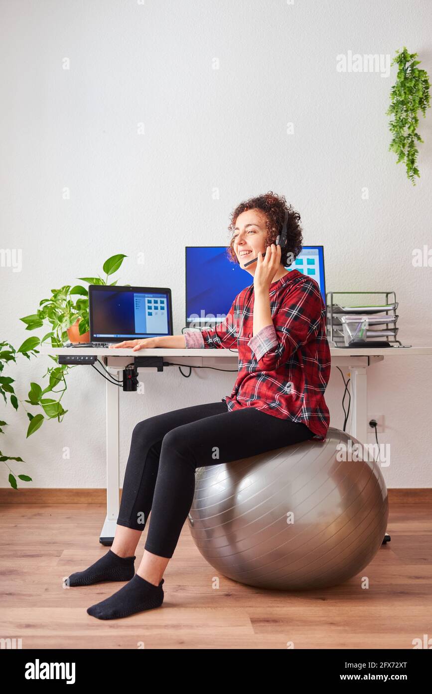 Woman teleworking sitting on a fitball in front of her desk talking on a headset Stock Photo