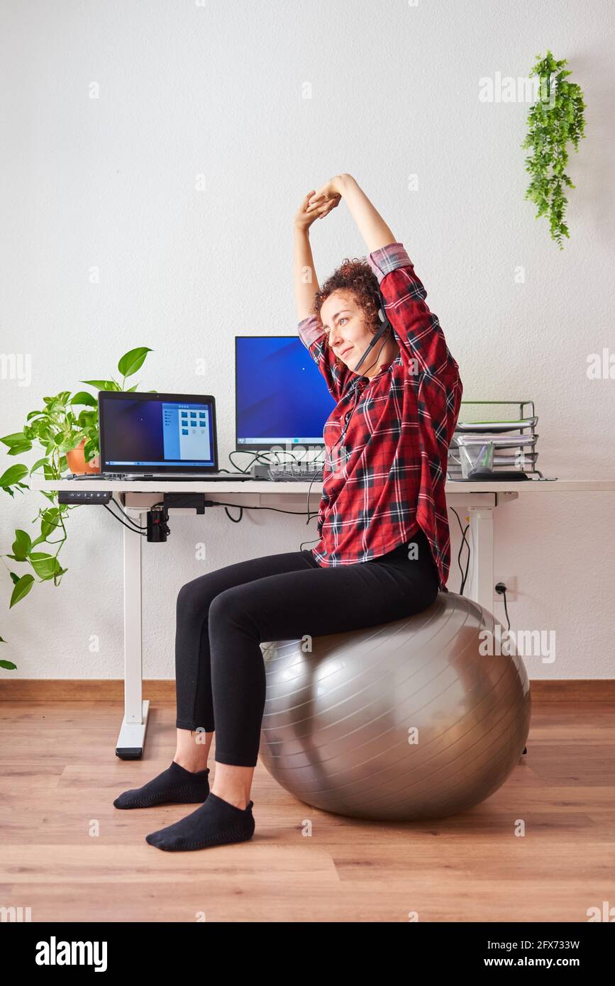 Woman stretches while working sitting on a fitball in front of her office desk Stock Photo
