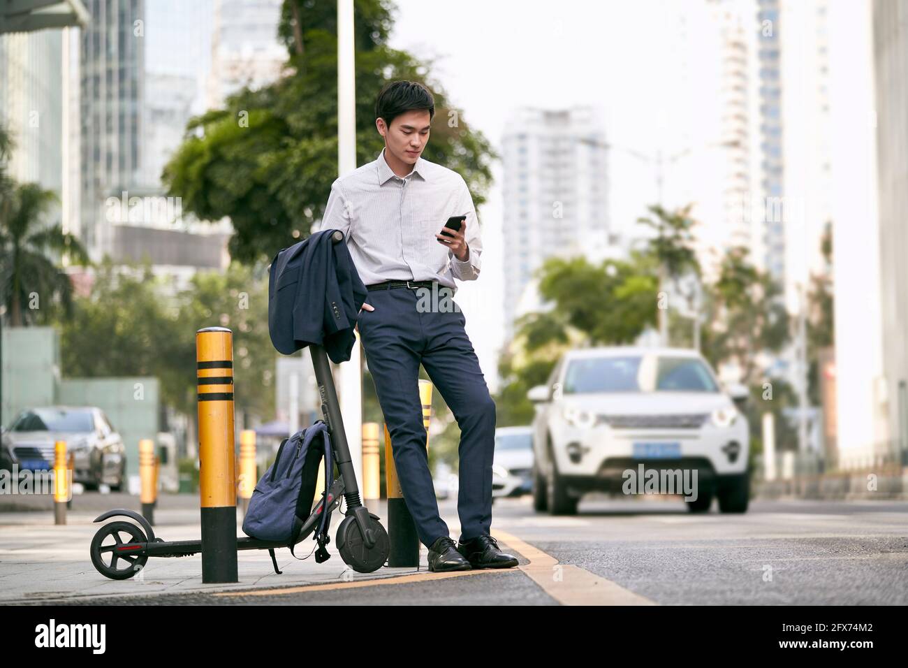 young asian businessman standing next to his electric scooter looking at cellphone on street in downtow of modern city Stock Photo