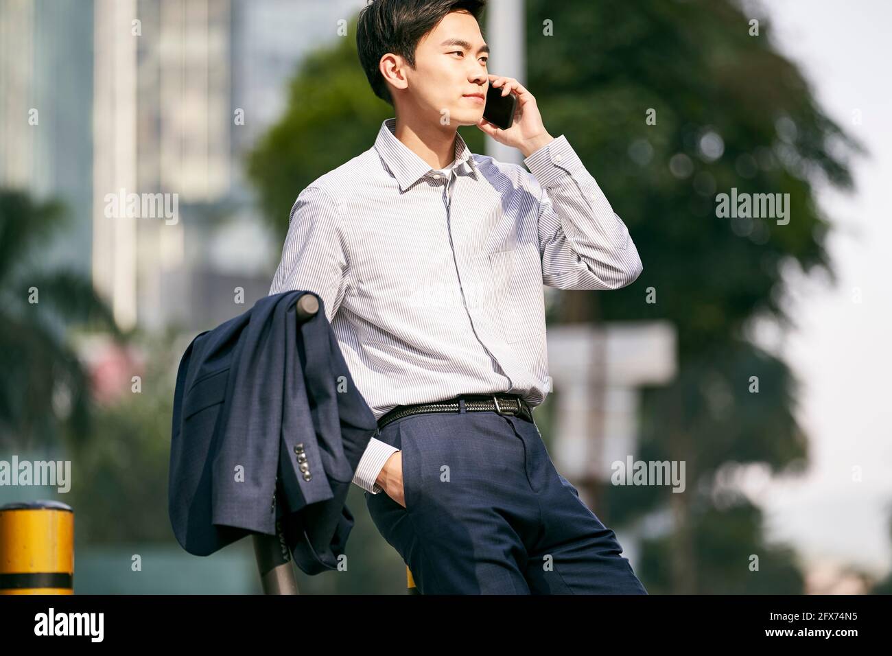 young asian businessman standing next to his electric scooter on street making a call using cellphone in downtow of modern city Stock Photo