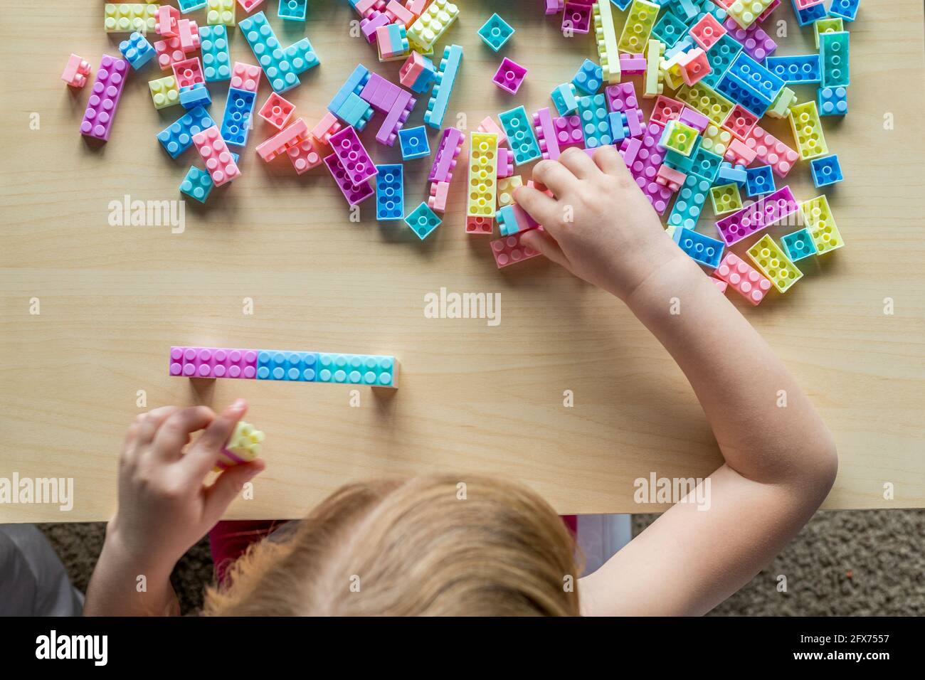 Little beautiful girl playing with toy plastic building blocks, sitting at the table. Small child busy with fun creative leisure activity. Development Stock Photo