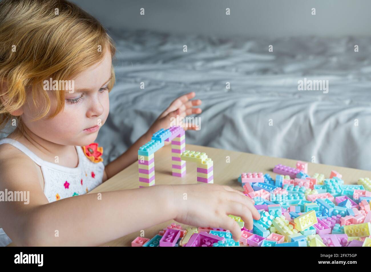 Little beautiful girl playing with toy plastic building blocks, sitting at the table. Small child busy with fun creative leisure activity. Development Stock Photo