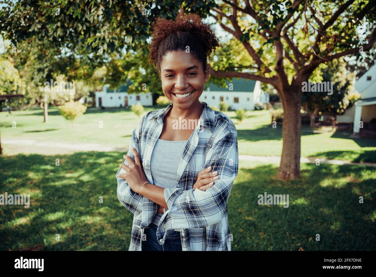 Mixed race female standing in farm village smiling with arms crossed in luscious green fields  Stock Photo