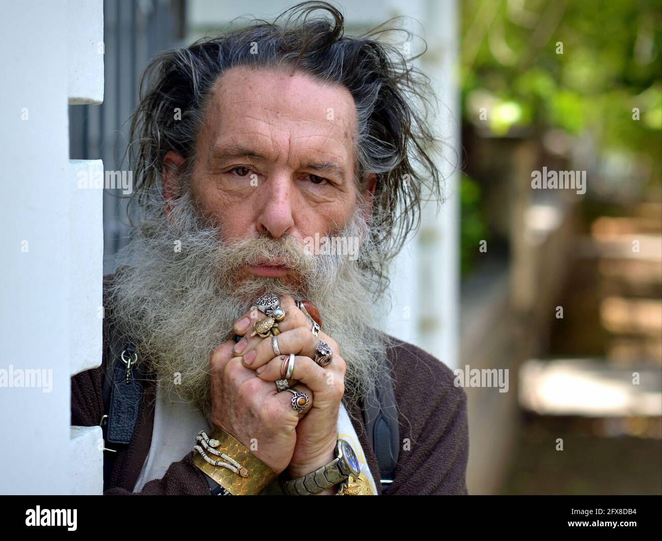 Old spiritual Caucasian wise man with thick beard and disheveled hair poses for the camera, clasps his hands and shows his many rings and bracelets. Stock Photo