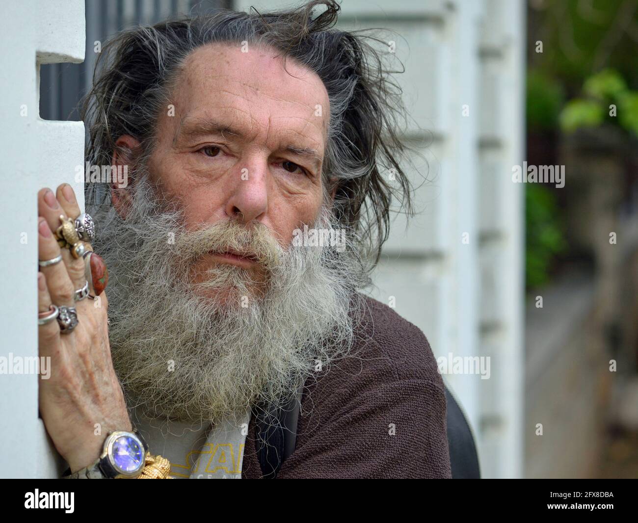Old emotional dignified Caucasian man with thick beard and disheveled hair hides behind a wall, looks at the viewer and shows the rings on his hand. Stock Photo