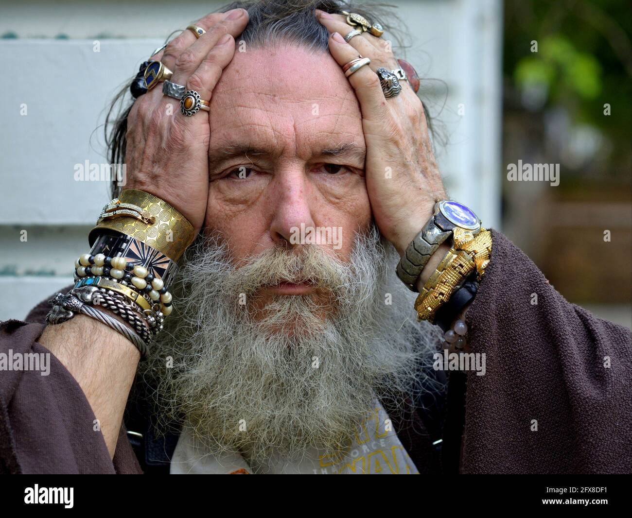Old esoteric Caucasian wise man with thick grey beard poses for the camera, holds his head in his hands, and shows his assorted rings and bracelets. Stock Photo