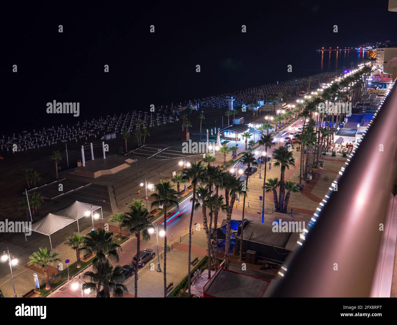 Top aerial view overlooking night lit Finikoudes Palm tree promenade, road with car lights, beach umbrellas near the Mediterranean sea and horizon. Stock Photo