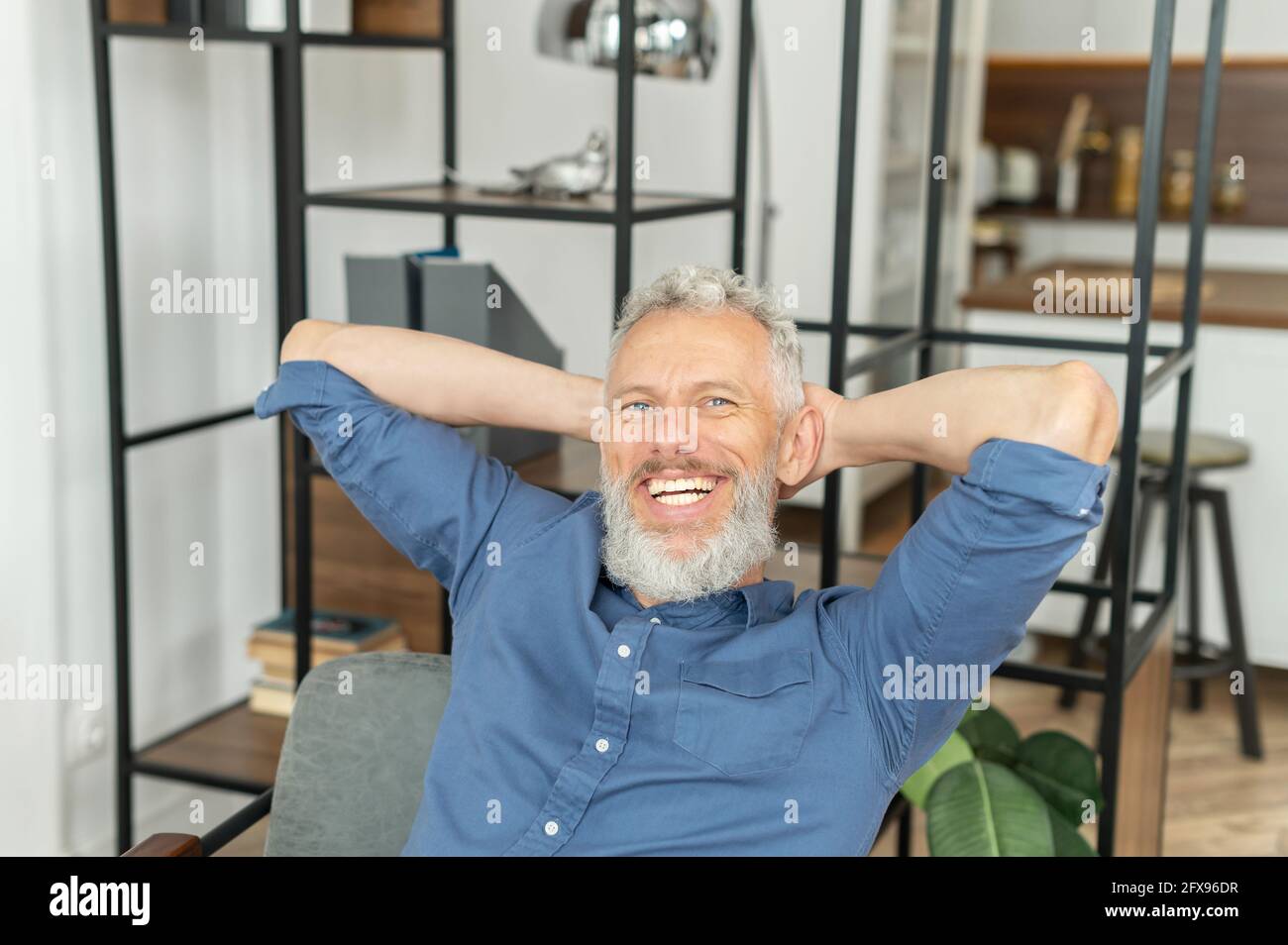 Relaxed cheerful senior grey-haired man resting in the office, mature charismatic businessman in smart casual shirt enjoying finish of good work day, day off starting, holding hands behind head Stock Photo