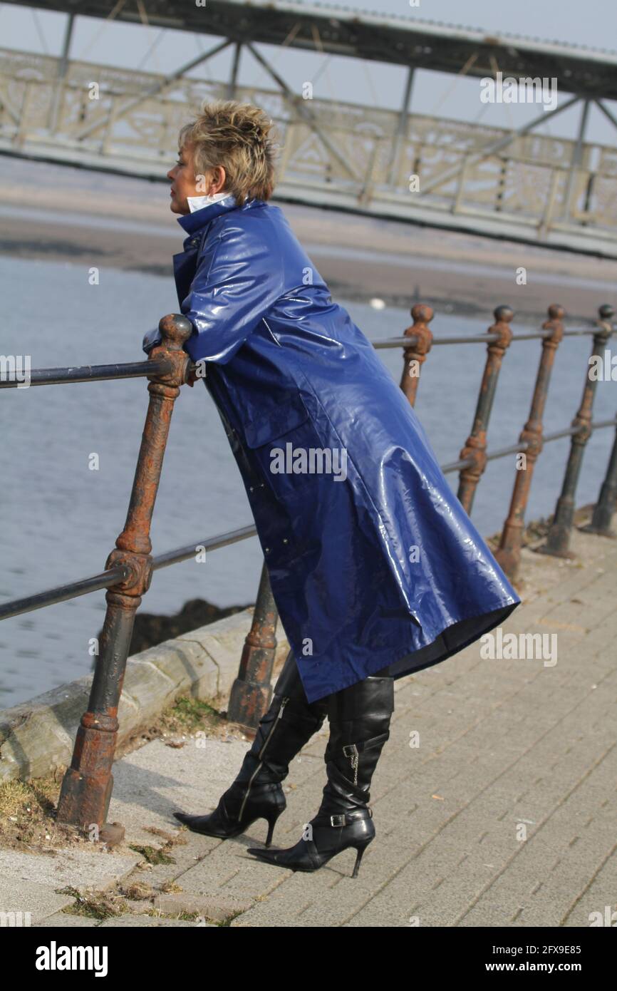 White caucasian middle aged woman with cropped spikey dyed hair wearing blue PVC raincoat leaning on railing at riverside looking for someone Stock Photo