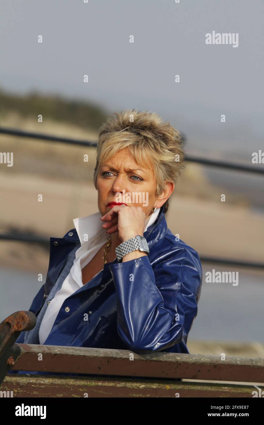 White caucasian middle aged woman with cropped spikey dyed hair wearing blue PVC raincoat sat on bench at river harbour location looking pensive, puzzled, lost side on to camera Stock Photo