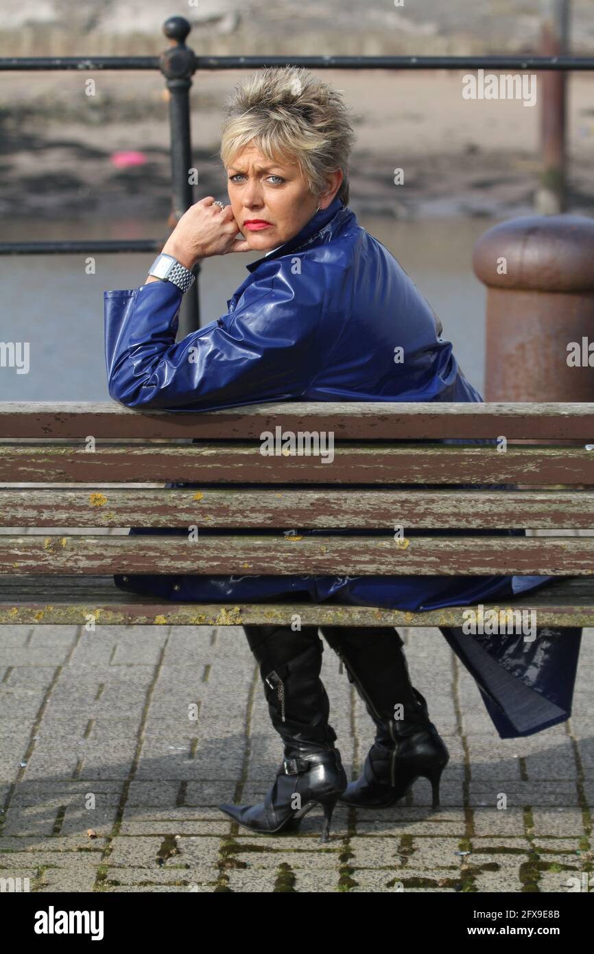 White caucasian middle aged woman with cropped spikey dyed hair wearing blue PVC raincoat sat on bench at river harbour location looking pensive, puzzled, lost side on to camera Stock Photo