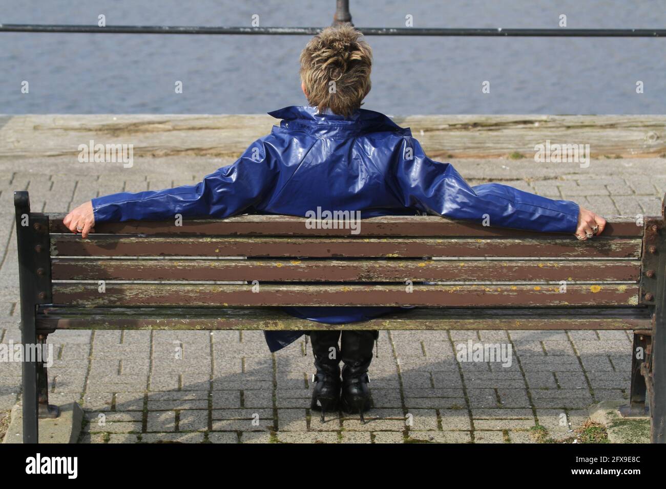 White caucasian middle aged woman with cropped spikey dyed hair wearing blue PVC raincoat sat on bench at river harbour location looking pensive, puzzled, lost , back to camera Stock Photo