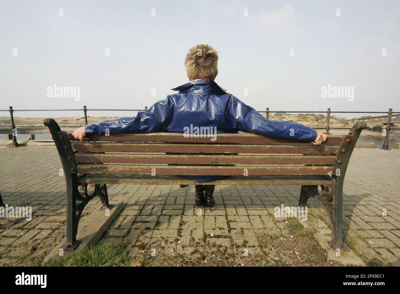 White caucasian middle aged woman with cropped spikey dyed hair wearing blue PVC raincoat sat on bench at river harbour location looking pensive, puzzled, lost , back to camera Stock Photo